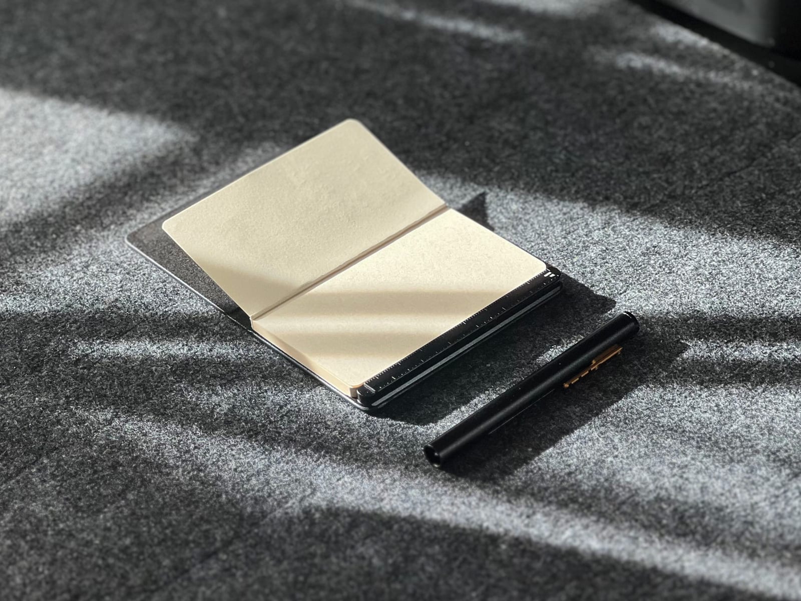 The noble. note lying open on grey felt in low sun, showing bone coloured paper and the black pen beside it